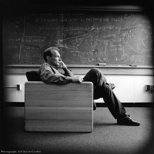 Photo of Tim Berners-Lee sitting in a chair. Behind him is a chalkboard coverd in mathematical equations.